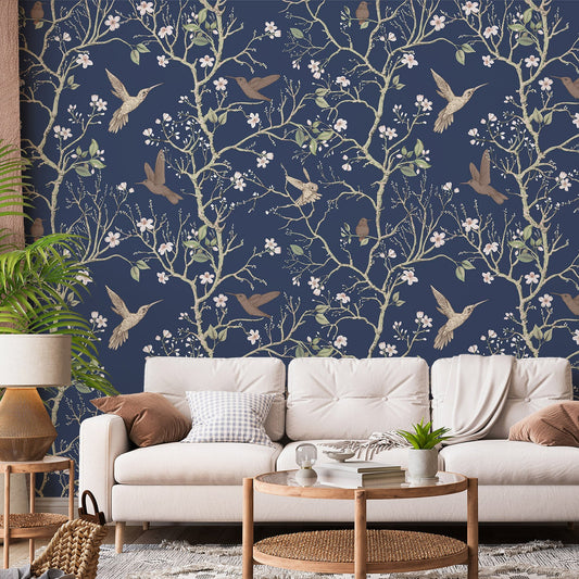 A living room with a cream sofa and a rattan coffee table showing 'Denora' which is a dark navy wallpaper featuring hand-drawn chinoiserie trees, birds and colourful flowers in an elegant repeating botanical pattern.