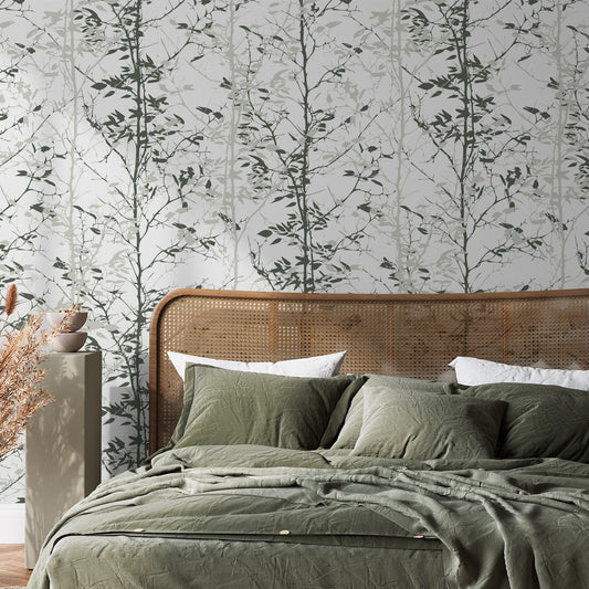 A bedroom with rattan furniture and green bedding showing 'Kiri' which is a green wallpaper featuring layered forest trees in varying shades of green with a hand-painted botanical landscape design.
