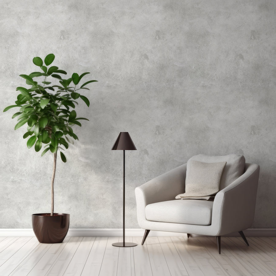 A living room with a grey sitting chair next to a standing lamp and leafy plant featuring the Ash wallpaper which is a grey concrete effect wallpaper with a detailed stone-like texture and soft tonal variations for an industrial look.