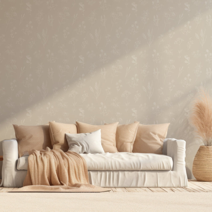 A living room with soft features such as a fabric sofa with scattered pillows and a blanket draped across it, next to a rattan plant pot with pampas grass. Showing 'Billie' which is Beige wallpaper featuring delicate white botanical outlines of flowers, leaves and plants in an intricate repeating pattern on a soft neutral background.