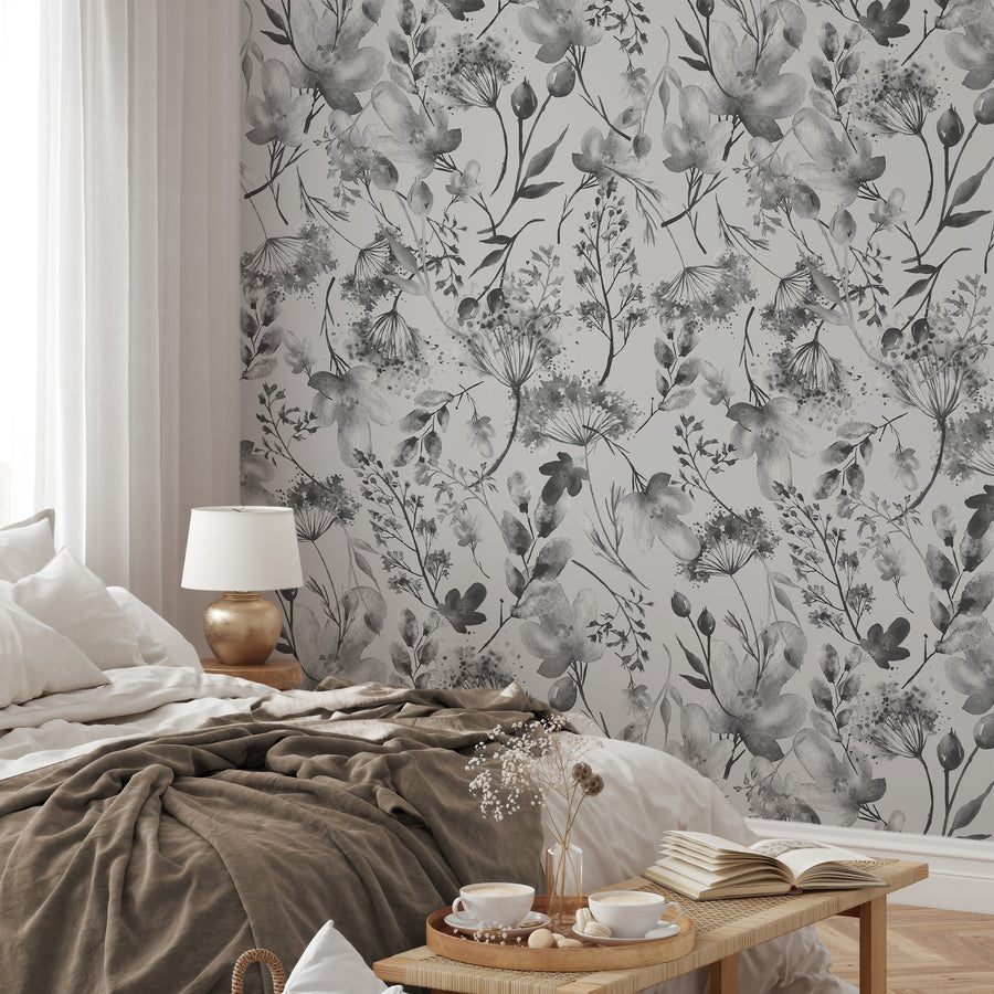 A relaxed styled bedroom showing the 'Matilda' which is a grey and white wallpaper featuring delicate watercolor floral illustrations with soft botanical blooms and leaves in a repeating pattern.