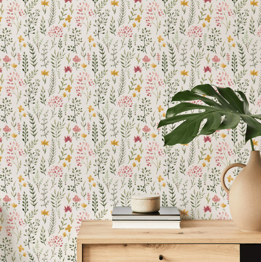 A living room with a side cupboard with a plant and books on top showing 'Mini Rosanna' which is a mini floral wallpaper featuring small watercolour flowers and leafy plants in soft green, yellow and pink tones on a beige background in a delicate repeating pattern.