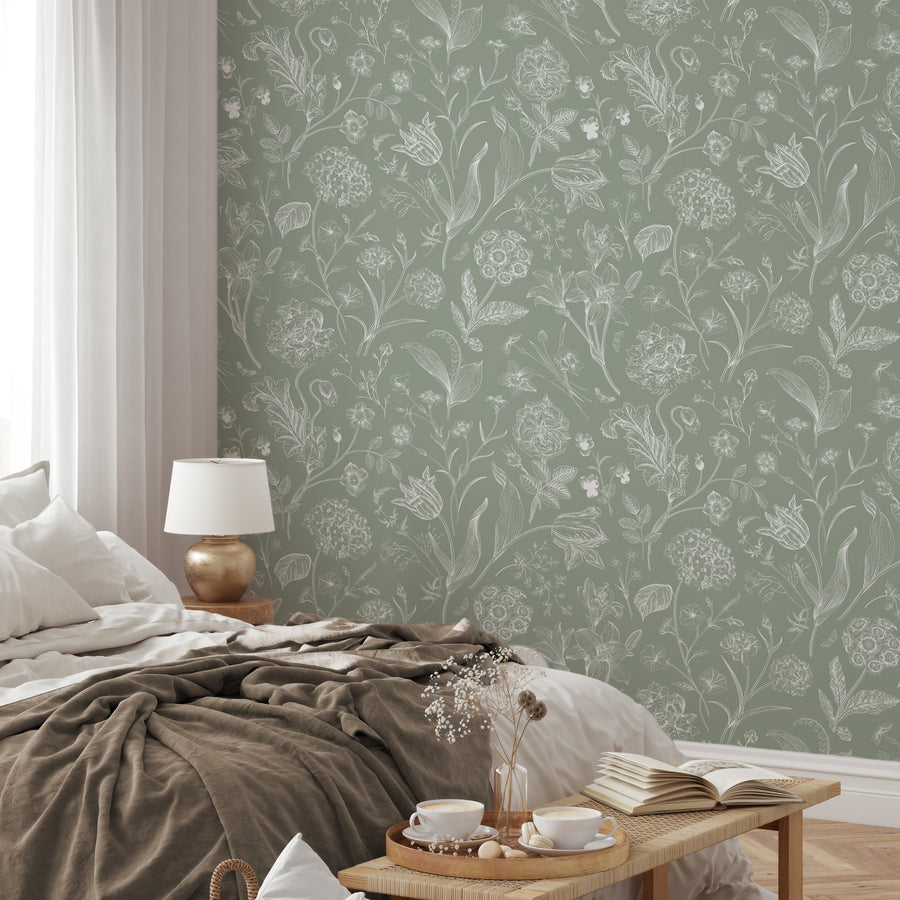 A relaxed styled bedroom showing 'Priscilla' which is a botanical wallpaper featuring delicate white floral and plant illustrations on a soft green background in a vintage-inspired repeating pattern.