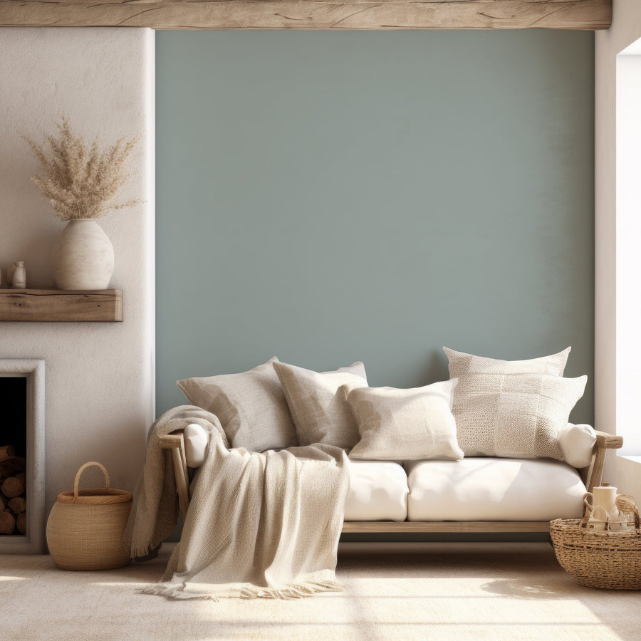 A coastal style living room with a cream sofa with pillows and blankets scattered across it with baskets surrounding and a fire place. This is showing 'Sarla' which is a slate grey wallpaper featuring a smooth solid colour finish with a subtle matte texture for a minimalist modern backdrop.