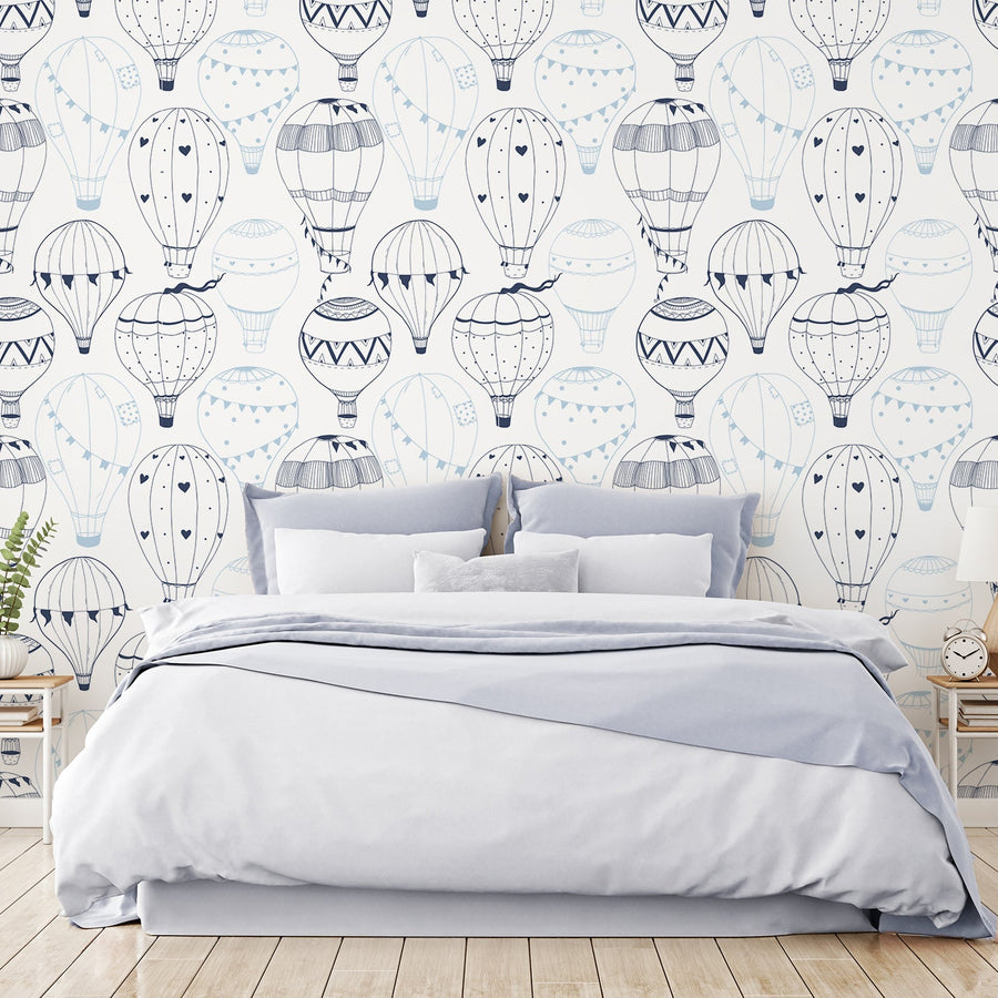 A bedroom showing a blue bed featuring 'Bellerose' which is a nursery wallpaper featuring dark and light blue illustrated hot air balloons floating across a white background in a charming repeating pattern.