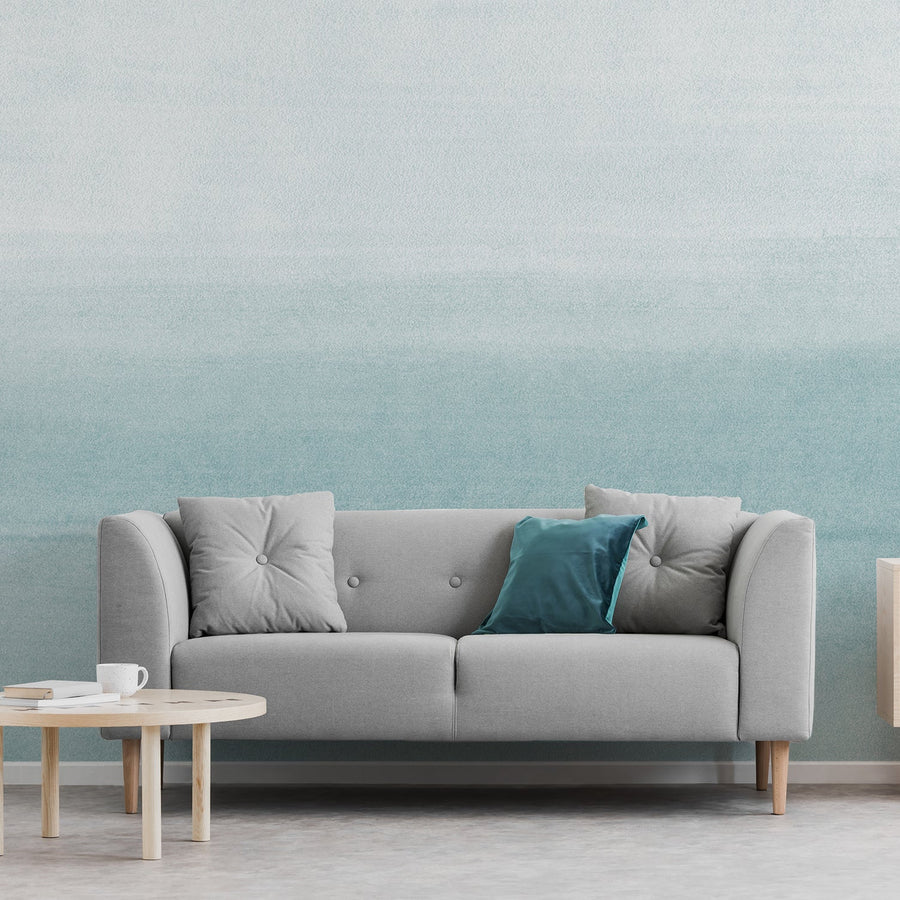 A living room with a two seater grey sofa featuring the Clara Teal wallpaper Teal wallpaper which is a soft watercolour wash effect with a subtle ombré gradient transitioning from deeper teal to lighter tones.