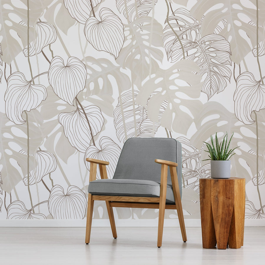 A living room with a grey chair next to an amber side table which features the Daphne wallpaper showing large monstera leaves with delicate line-art detailing in a soft repeating botanical pattern in neutral colours.