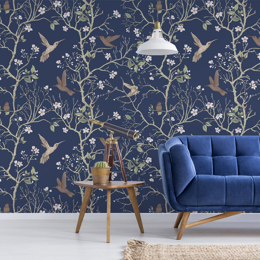 A living room with a blue tufted sofa and a small side table showing 'Denora' which is a dark navy wallpaper featuring hand-drawn chinoiserie trees, birds and colourful flowers in an elegant repeating botanical pattern.