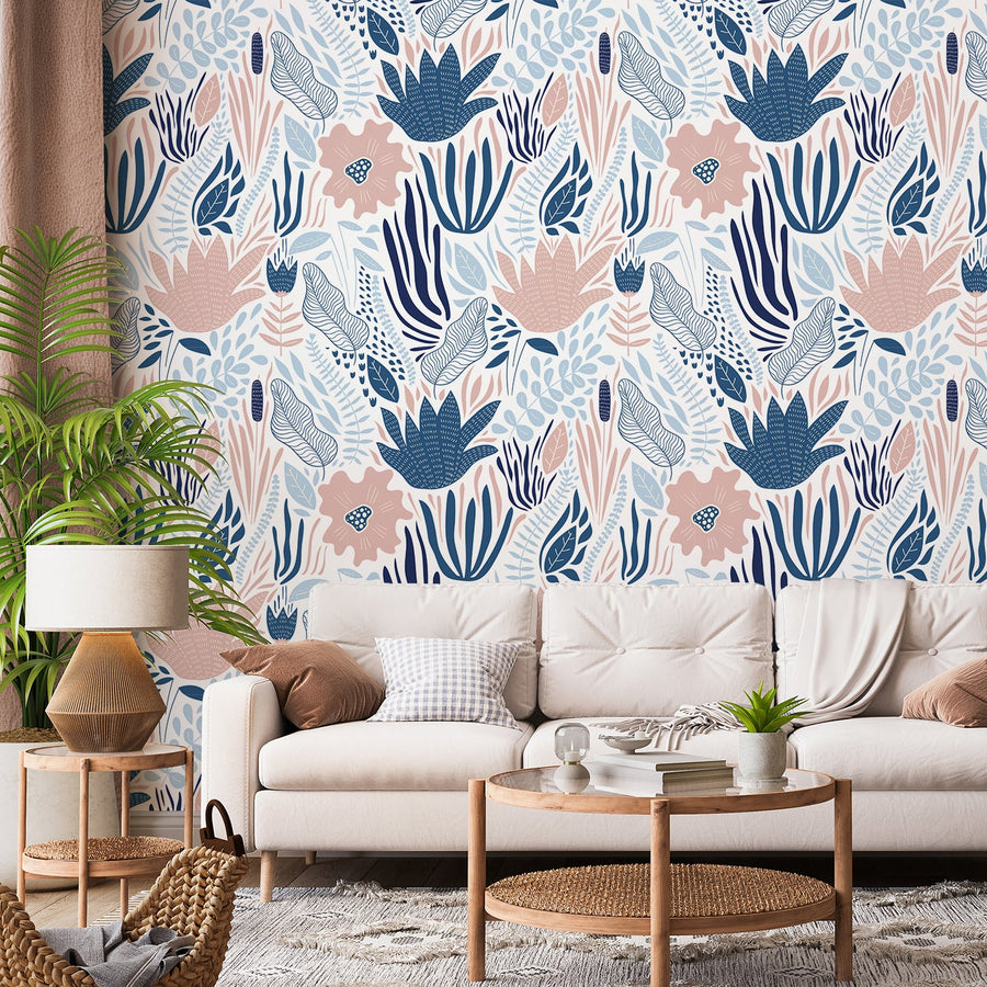 A living room with a cream sofa surrounded by wooden rattan furniture and a large leaf plant showing 'Eloise' which is a tropical wallpaper featuring blue and pink botanical plant silhouettes with leafy foliage on a soft white background in a vibrant repeating pattern.