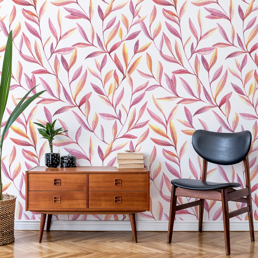 A living room with a leather black chair, amber wood chest of drawers with leafy green plants featuring 'Perry' which is a botanical wallpaper featuring pink and orange watercolour leaves on a crisp white background in a soft contemporary repeating foliage pattern.